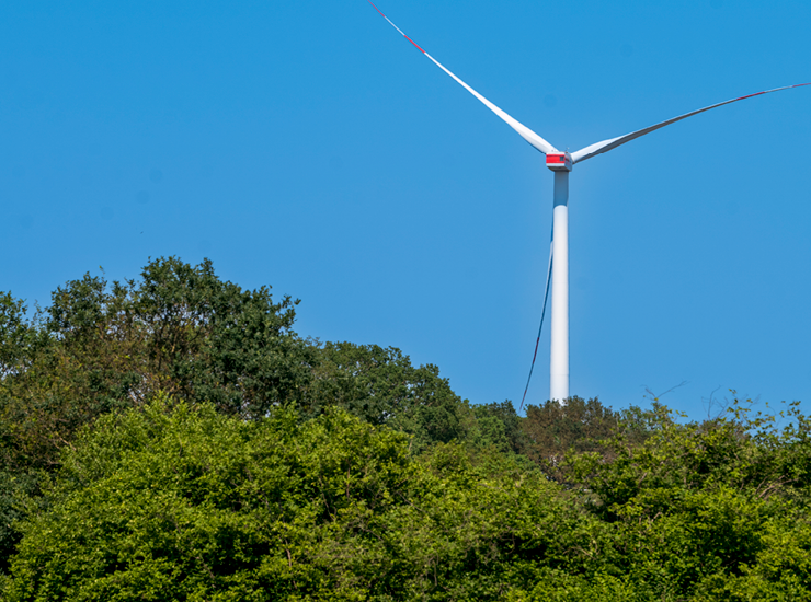 Windrad vor blauem Himmel mit einem grünen Wald im Vordergrund
