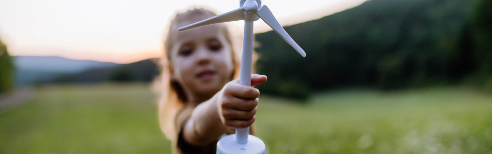 Kleines Mädchen hält Modell einer Windenergieanlage. Wiese im Hintergrund.